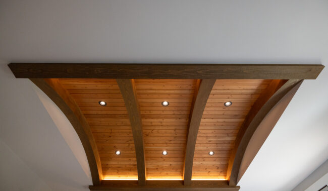 Wooden vaulted ceiling feature with curved beams, recessed lights, and warm accent lighting, set against a white ceiling background.