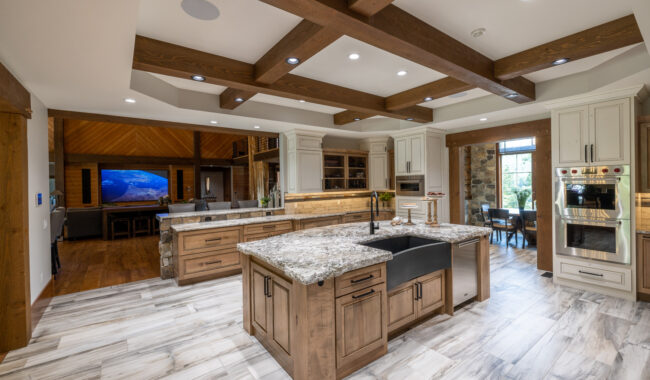 Spacious modern kitchen with wooden beams, marble countertops, a large island with a black sink, light cabinets, and stainless steel appliances; open to a dining and living area with wood floors and a TV.