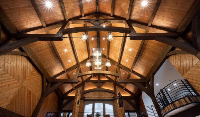 A view of a vaulted wooden ceiling with exposed beams, modern chandeliers, and spotlights. Below, a balcony with black railings and two tall wooden pillars enhance the rustic yet elegant interior design.