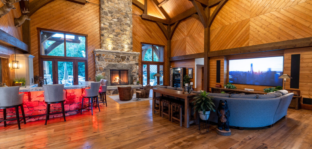 Spacious rustic living room with vaulted wood ceilings, exposed beams, a stone fireplace, a bar with red lighting, large windows, and modern furniture, including a gray sofa facing a large TV.