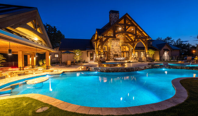 A luxurious house with warm lights stands behind a large, illuminated swimming pool at dusk. The pool features a hot tub and stone accents, surrounded by outdoor seating and a covered patio area.