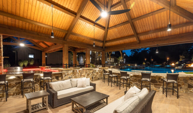 Covered outdoor lounge with wicker sofas and armchairs, stone bar seating, and wooden ceiling with exposed beams. The space overlooks a lit swimming pool at dusk, creating a warm, inviting atmosphere.