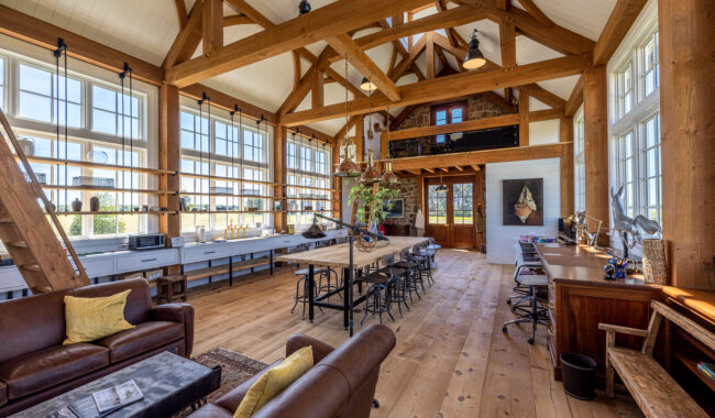Spacious, sunlit room with high wooden beams, large windows, a long dining table, leather sofas, and multiple workstations. The decor combines rustic and modern elements, creating a cozy and inviting atmosphere.