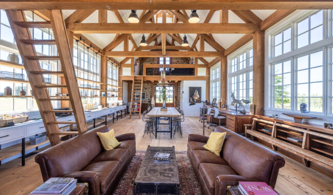 Spacious, sunlit room with exposed wooden beams, large windows, two brown leather sofas, a long wooden dining table, bookshelves, and various decorative objects; rustic and cozy interior design.