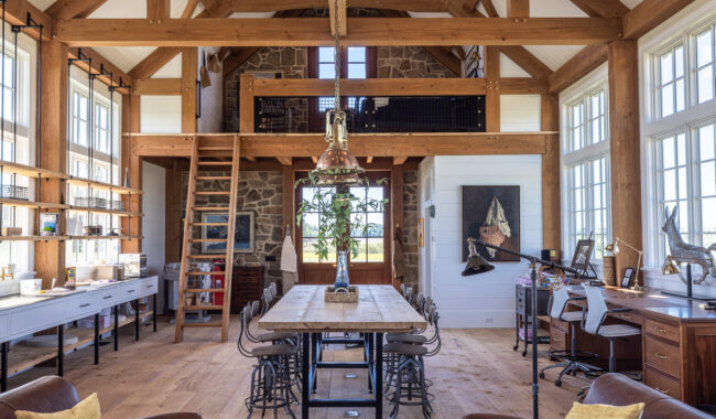 A spacious, sunlit room with exposed wooden beams, a long wooden dining table and chairs, large windows, bookshelves on the left, desks on the right, and a loft area above with a stone accent wall.