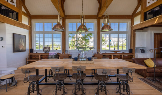 Spacious, rustic dining area with a long wooden table, six metal bar stools, large windows letting in natural light, wooden beams on the ceiling, and pendant lights hanging above the table.