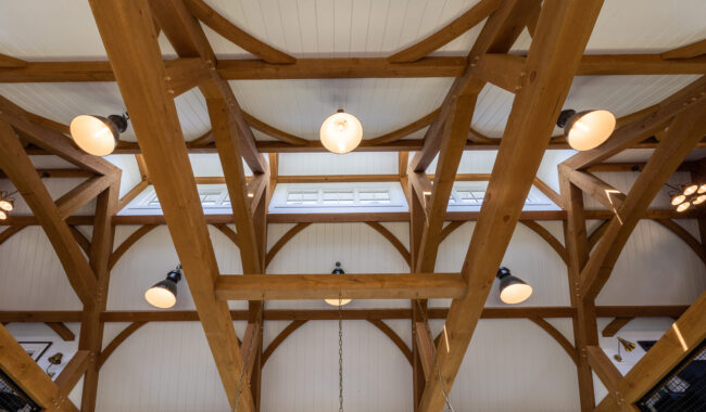 A wooden beam ceiling with white panels, exposed timber trusses, and hanging round light fixtures, viewed from below. Rectangular windows near the ceiling let in natural light.