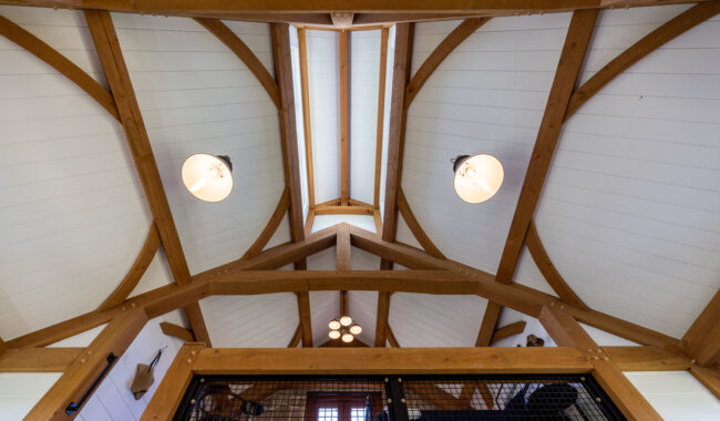 Upward view of a vaulted ceiling with exposed wooden beams, white paneling, three round light fixtures, and a balcony railing with a mesh guard below.