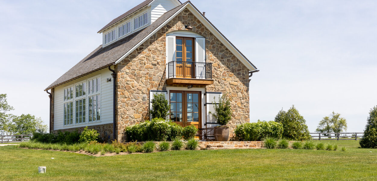 A charming stone house with large windows, a small balcony above the front door, and a well-manicured lawn surrounded by green bushes and trees under a partly cloudy sky.