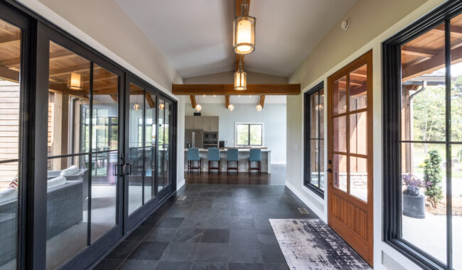 A bright hallway with slate tile flooring leads to a modern kitchen with blue barstools. Large windows and glass doors line the hallway, offering views of a sunroom and the outdoors. Light fixtures hang from a vaulted ceiling.