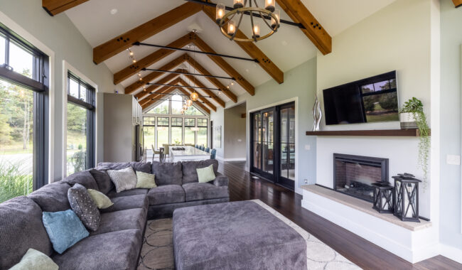 A modern living room with vaulted wood beam ceilings, large windows, a gray sectional sofa, a fireplace with a TV above it, dark wood floors, and a dining area in the background.