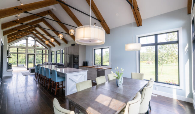 A bright, modern kitchen and dining area with wooden beams, a long island with blue stools, a large dining table with cream chairs, pendant lights, and large windows overlooking a green outdoor landscape.