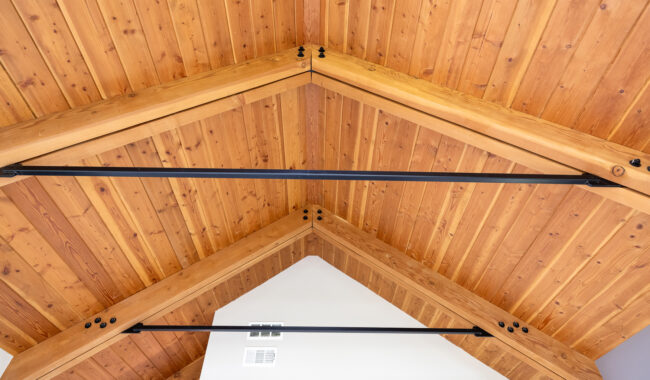 Angled view of a vaulted wooden ceiling with exposed beams and metal support rods, showcasing natural wood grain and a modern architectural design. White walls are visible at the peak.