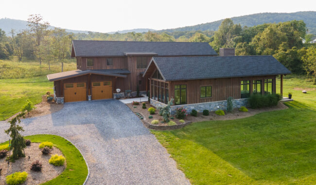 A modern two-story wooden house with a gravel driveway, landscaped garden, and large windows, set on a spacious green lawn with trees and hills in the background.