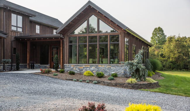 A modern house with large windows, stone accents, and wood siding, surrounded by landscaped shrubs, gravel driveway, and a green lawn with trees in the background.