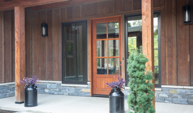 A wooden front door with glass panels is framed by two large windows. Two black planters with purple plants and a small evergreen shrub sit on the porch, which features wood columns and stone accents.