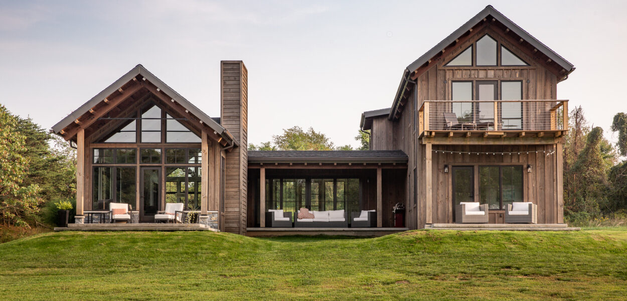 Modern two-story wooden house with large windows, two connected sections, a balcony, and outdoor seating on a spacious green lawn under a clear sky.