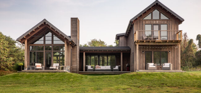 Modern two-story wooden house with large windows, two connected sections, a balcony, and outdoor seating on a spacious green lawn under a clear sky.