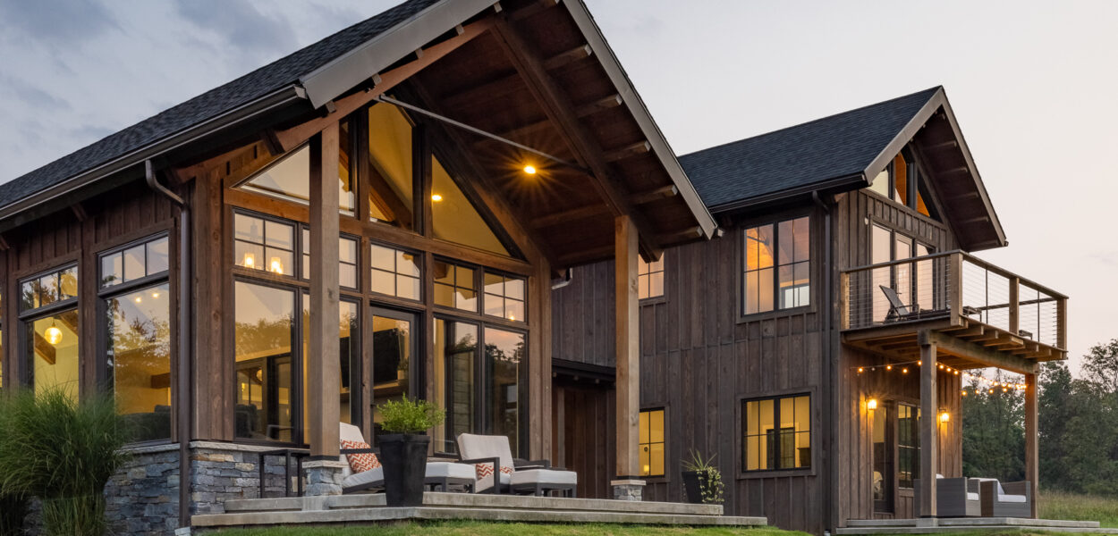 Modern two-story house with large windows, wooden siding, and stone accents. Outdoor seating on patio and balcony overlooks a green lawn at sunset. Warm interior lights are visible through the windows.