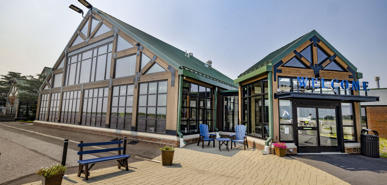A modern building with large glass windows, green peaked roofs, and a WELCOME sign above the entrance. Blue chairs and planters decorate the paved area outside.