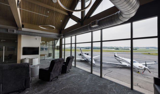 Modern airport lounge with large windows overlooking a runway, where two small private jets are parked. The lounge has comfortable black chairs, a TV, and visible exposed ducts and wooden ceiling beams.