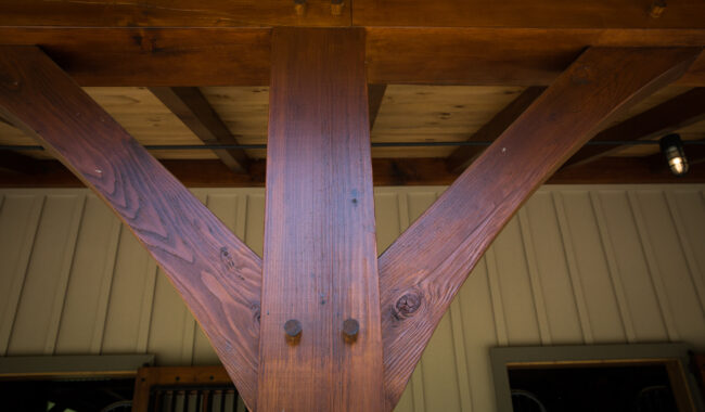 A close-up view of a wooden beam structure supporting a roof, showing two angled support beams meeting a vertical post, secured by large bolts. The background includes vertical wood siding and part of a window.