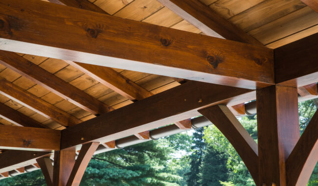 Close-up view of a wooden pergola roof with exposed beams, showcasing smooth, polished wood and sturdy construction. Green trees and foliage are visible in the background.