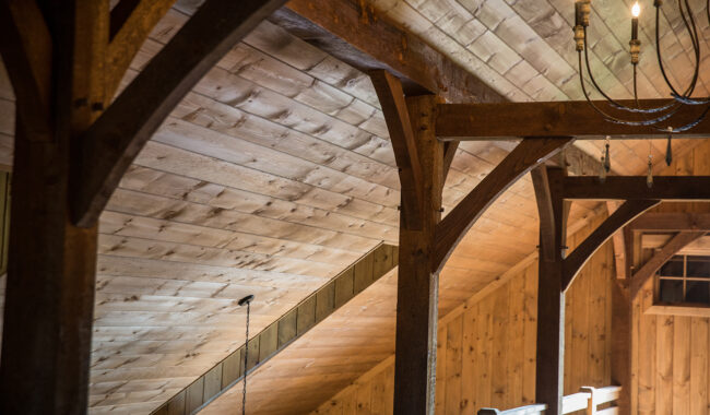 Wooden beams and planks form the slanted ceiling and upper walls of a rustic interior, illuminated by natural light and a metal chandelier, creating a warm and inviting atmosphere.