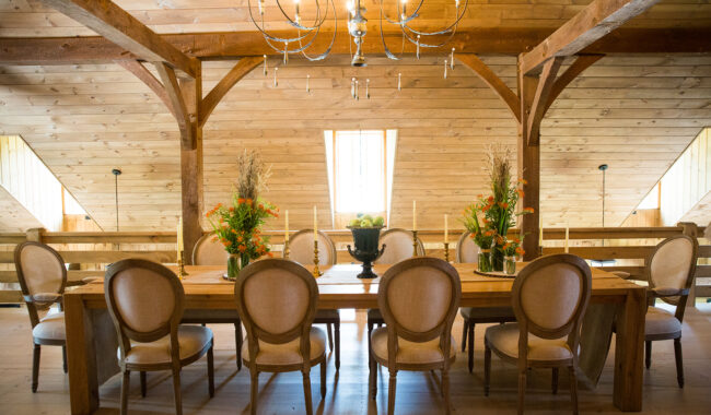 A rustic dining room with a long wooden table, ten upholstered chairs, floral centerpieces, candles, a black urn, and a chandelier hanging from a sloped, wood-paneled ceiling with exposed beams and windows.
