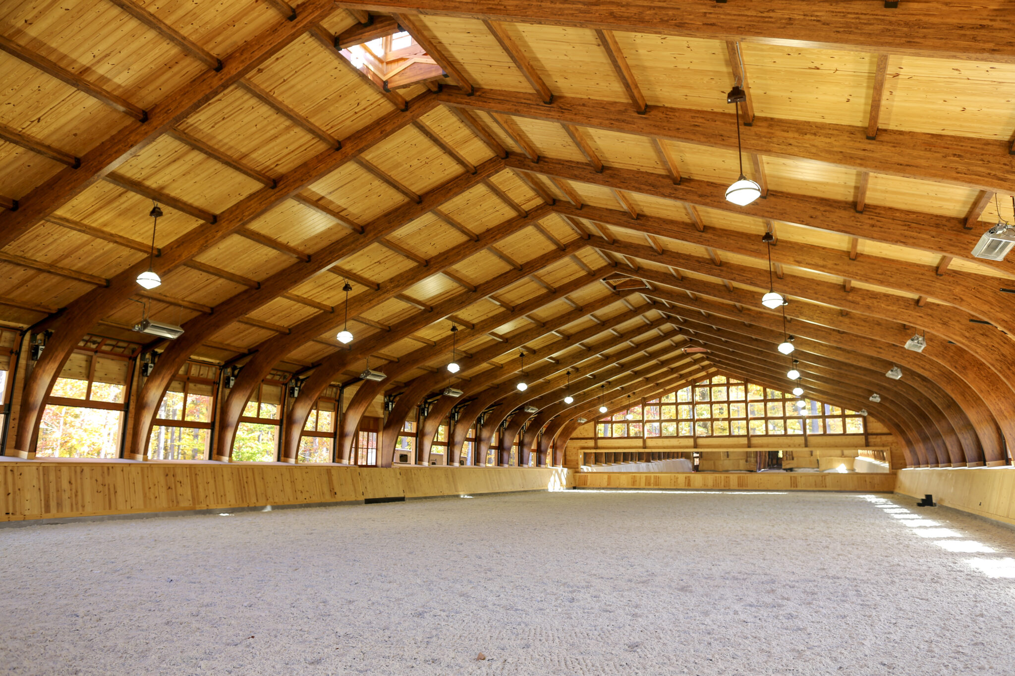 A spacious indoor riding arena, featured in our timber frame project gallery, boasts a high, arched wooden ceiling, large side windows, and sand-covered ground, all brightly lit by natural and overhead lights.