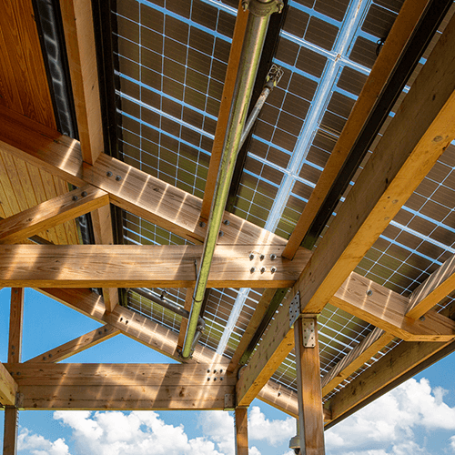 View from below of a wooden pergola structure with solar panels integrated into the roof, set against a blue sky with scattered white clouds.
