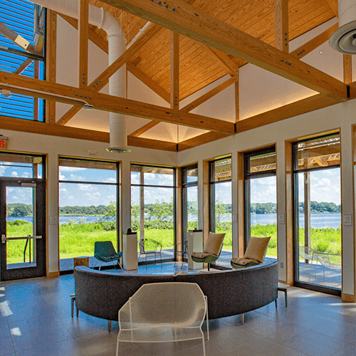 Modern room with large windows, wooden ceiling beams, and contemporary chairs. The space is bright with natural light and offers views of green grass and a lake outside.