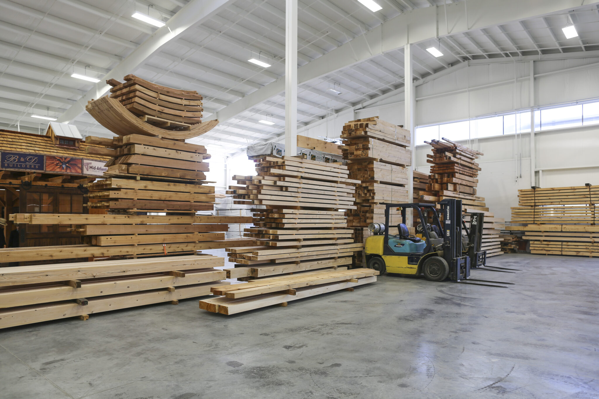 Stacks of large wooden beams and planks are organized inside a spacious, well-lit warehouse. Nearby, a yellow and green forklift is parked by the wood piles—perfect inspiration for any timber frame project gallery.