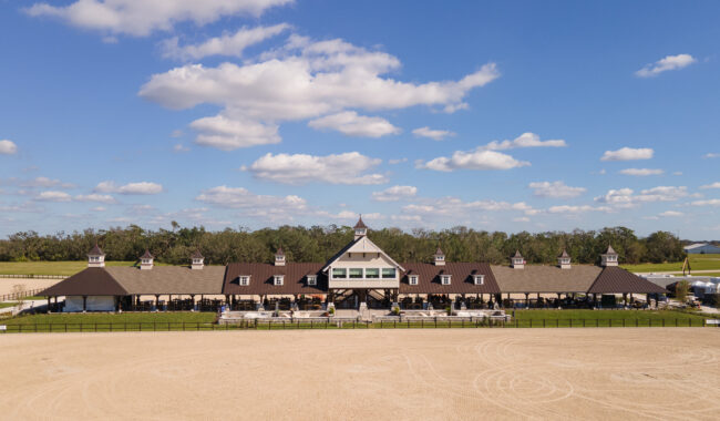 A large, elegant equestrian barn with cupolas stands in the center of a sandy riding arena under a blue sky with scattered clouds, surrounded by green fields and trees.