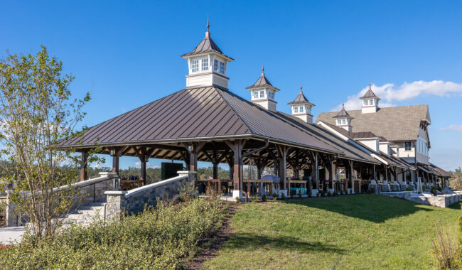 Large open-air pavilion with a dark metal roof and several cupolas, set amid green grass and landscaping, under a clear blue sky. The structure features wooden beams and stone accents.