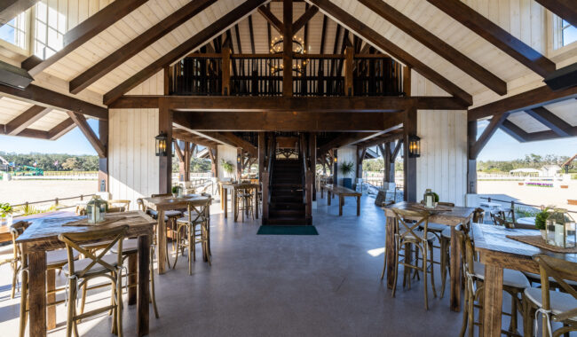 Spacious, rustic dining area with wooden tables and chairs, high vaulted ceiling with exposed beams, a central staircase, large windows, and views of an outdoor area. Natural light fills the bright, open interior.