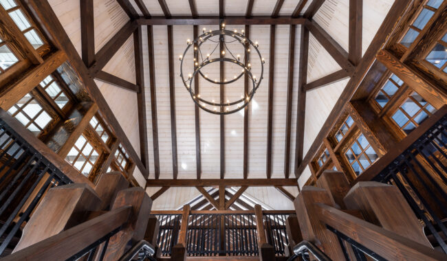 View looking up at a high ceiling with exposed wooden beams, a large circular chandelier, and surrounding wooden staircases and railings with windows on both sides letting in natural light.