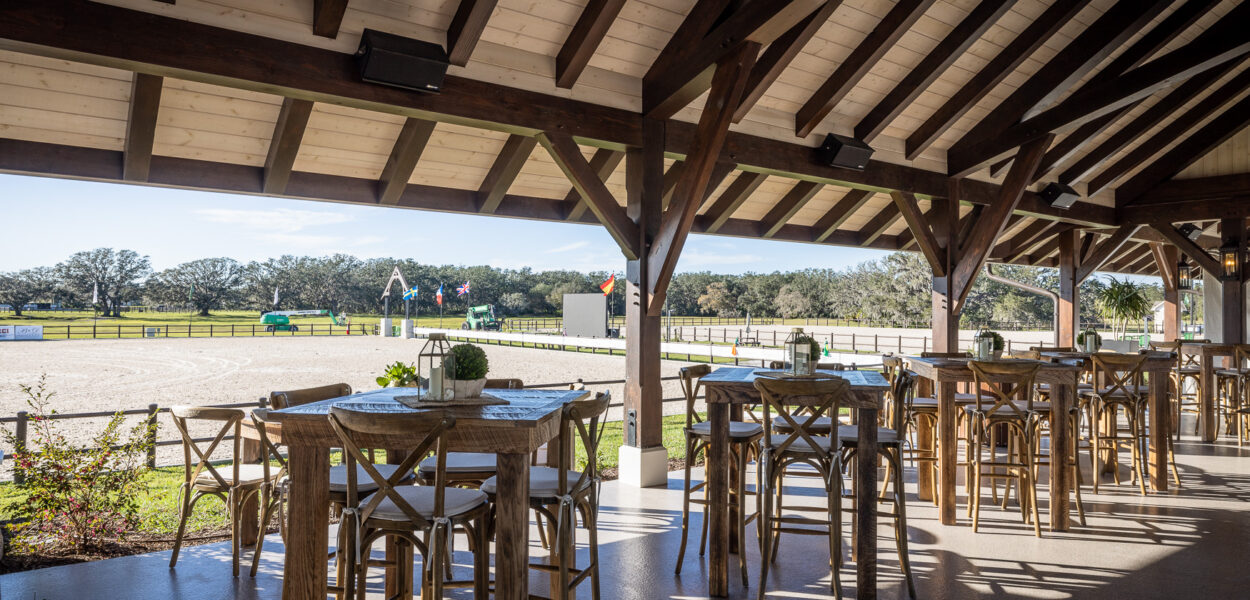 Open-air pavilion with wooden tables and chairs overlooking a fenced outdoor arena and green landscape under a clear sky. Small potted plants decorate the tables, and sunlight streams in from the side.