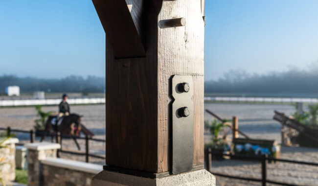 A close-up of a wooden post with metal brackets in the foreground, with an outdoor equestrian arena and a rider on a horse blurred in the background.