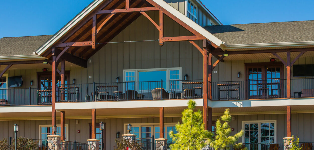 A large, two-story lodge-style building with wooden beams, balconies, and stone accents, set against a clear blue sky. The foreground features manicured grass, a small flag, and green landscaping.