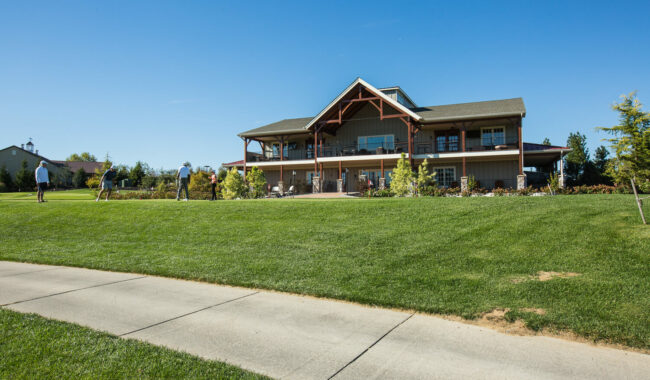 A group of people play golf on a green lawn in front of a large, two-story clubhouse with a wooden balcony, under a clear blue sky.