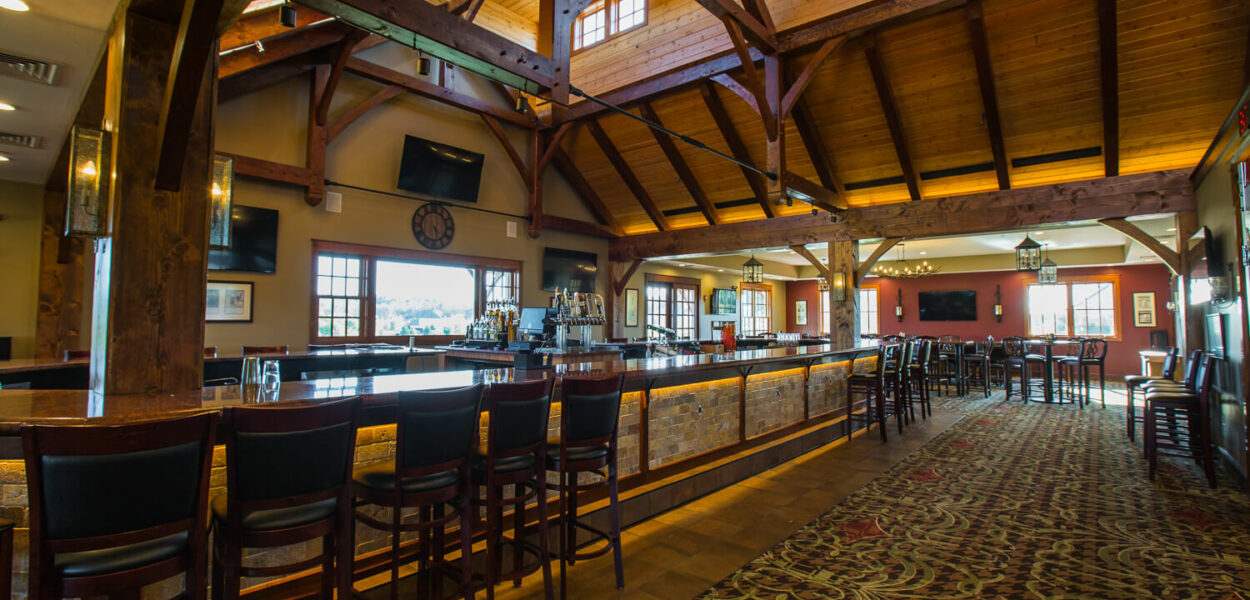 A spacious bar and restaurant interior with high wooden beam ceilings, a long bar lined with chairs, large windows, carpeted flooring, and tables with chairs in the background. Warm lighting creates a cozy atmosphere.