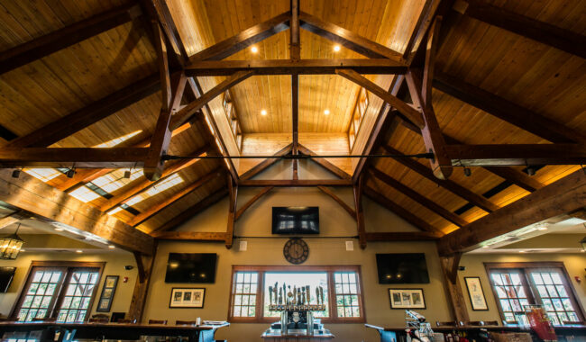 A spacious bar with a vaulted wooden ceiling, exposed beams, and large windows. Warm lighting highlights the rustic décor, wall-mounted TVs, framed art, and a central bar with taps.