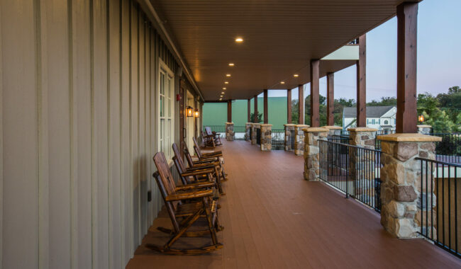 A long, covered porch with wooden rocking chairs lined up along the wall, stone columns, and metal railings overlooking a neighborhood with houses and trees in the background.