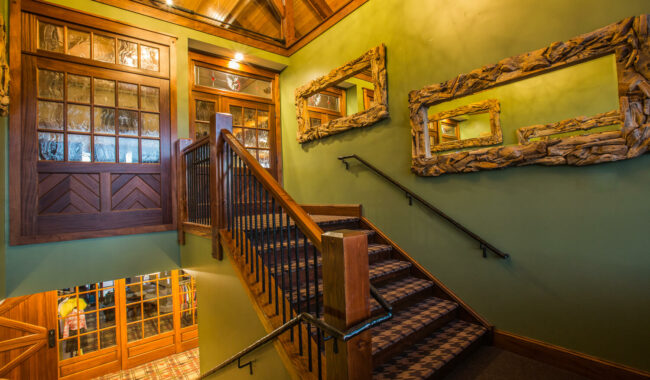A wooden staircase with patterned carpet leads to an upper floor. The green walls feature two large, rustic mirrors with wooden frames. Glass doors and windows let in warm light, creating a cozy, inviting atmosphere.