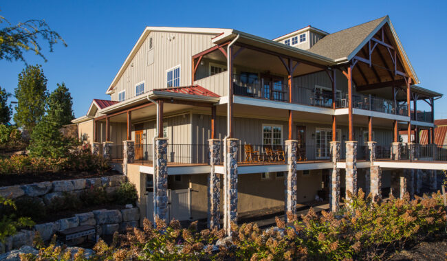 A large, modern two-story house with stone pillars, wood accents, multiple balconies, and large windows, surrounded by landscaping and greenery under a clear blue sky.