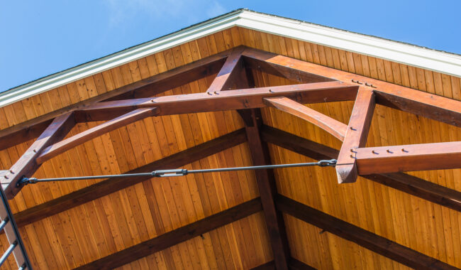 Close-up view of a wooden roof with exposed beams and trusses against a clear blue sky, showing the structure and natural wood texture.