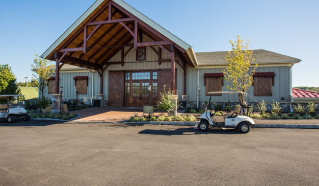 A rustic building with a wooden entrance and large barn-style doors, set on a paved area. Two golf carts are parked in front, and small trees and plants line the walkway.