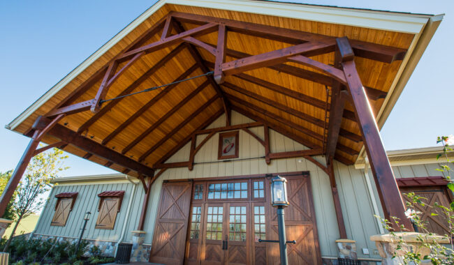 A rustic building with a high, wooden A-frame entrance, large double barn doors, and stone details, viewed from below on a sunny day. A lantern stands near the doorway, and landscaping surrounds the entrance.