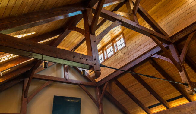 Exposed wooden beams and trusses create a rustic ceiling in a bright room with a mounted TV, beige walls, and windows letting in natural light.